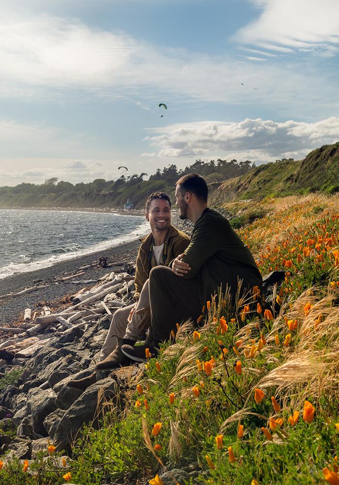 A male couple sits amidst wildflowers along the coast of Victoria, BC while paragliders sail through the sky behind